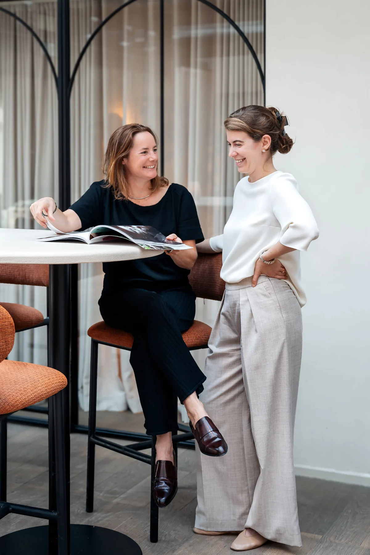Alexia Hoste en Charlotte Knockaert bekijken lachend samen een tijdschrift aan een ronde statafel, voor een raam met een boog.